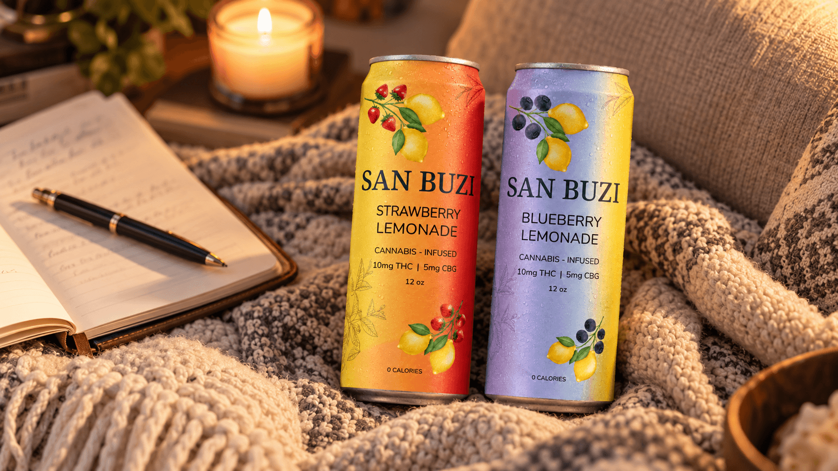 San Buzi Strawberry Lemonade and Blueberry Lemonade cans on a cozy blanket next to an open journal and a lit candle