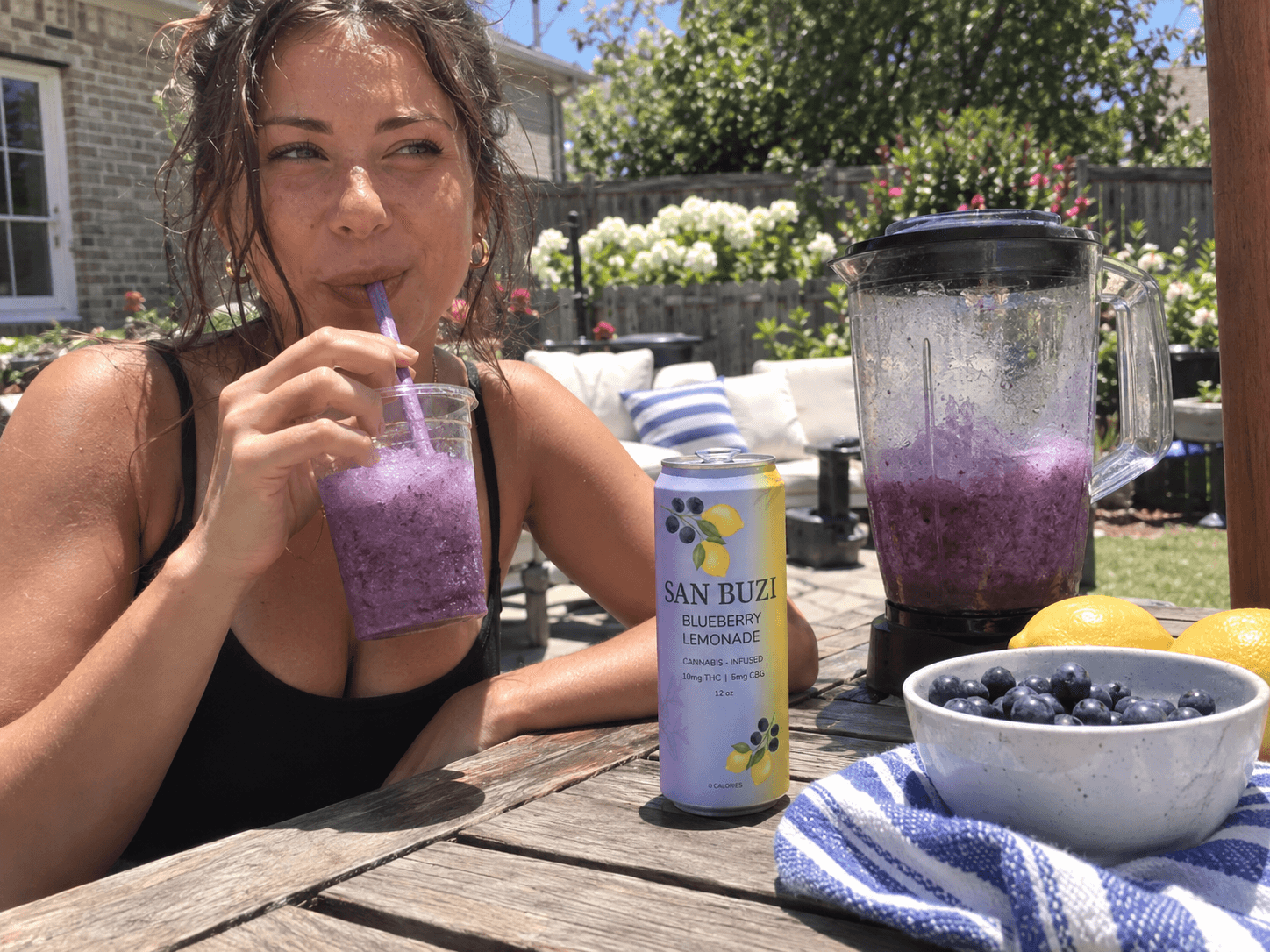 A woman drinking a blueberry San Buzi slushie through a straw at an outdoor table in bright sun, with a blender and bowl of blueberries